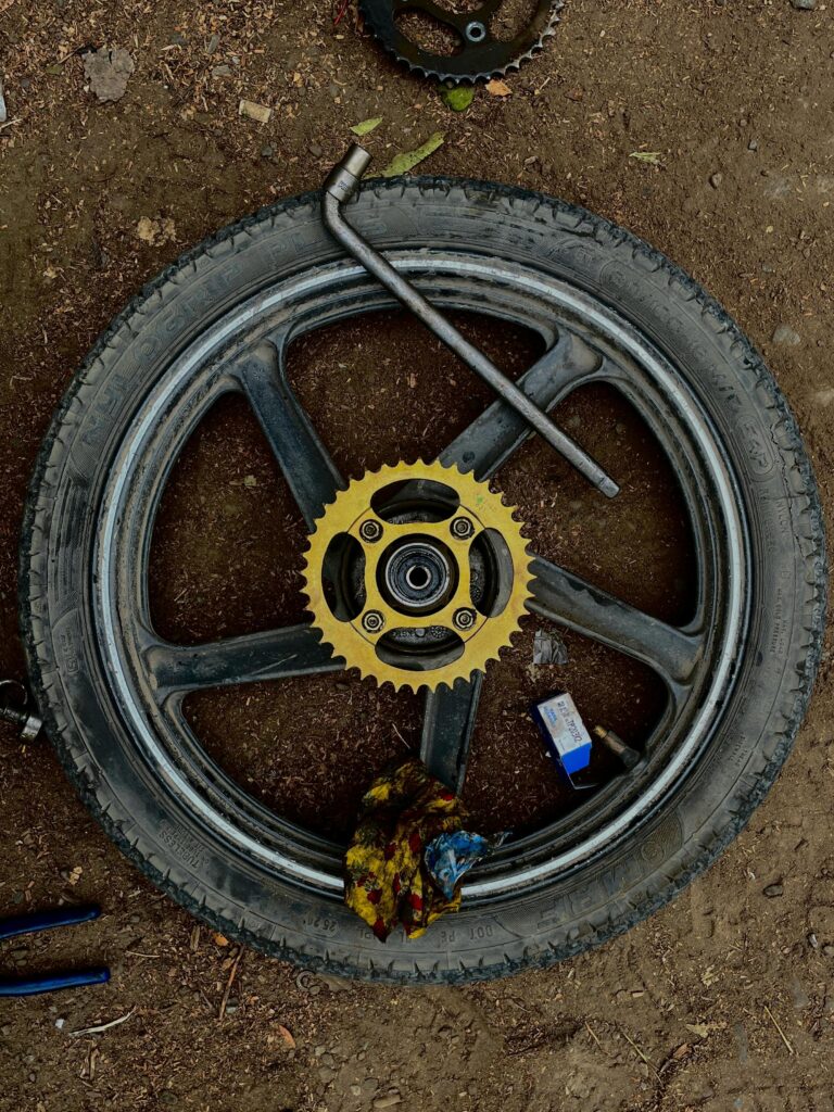 Motorcycle wheel with sprocket and tools on dirt ground in DIY repair setup.